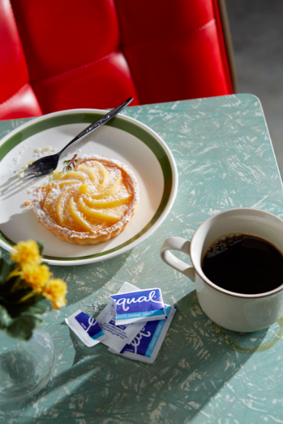 Overhead shot of a lemon tart, sugar packets, a cup of coffee, and yellow flowers on a speckled blue table with a red bench.