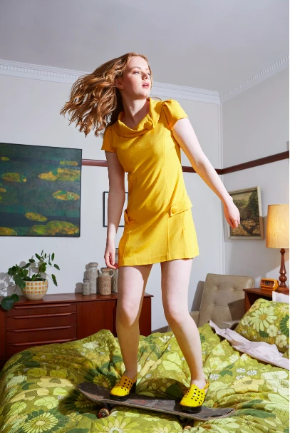 Young woman in a yellow dress balances on a skateboard atop a floral bedspread in a sunlit bedroom with vintage decor and a painting.