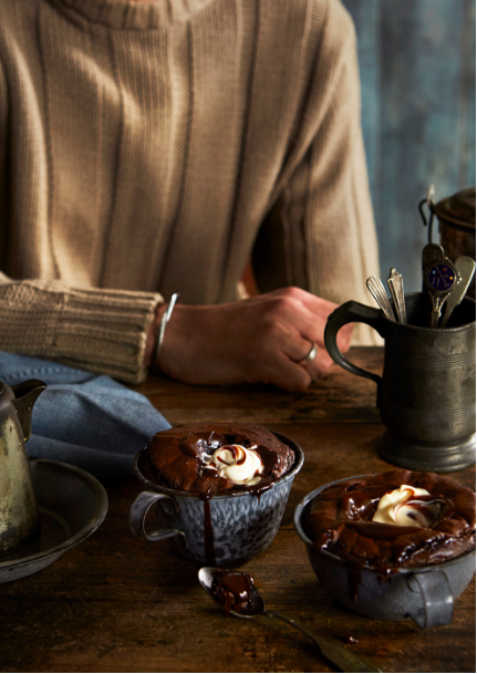 Eye-level shot of two chocolate mug cakes topped with whipped cream served on a rustic wooden table with a person in background.