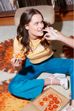 A woman in a yellow t-shirt and blue pants eating pizza on a patterned rug indoors, photographed by Heather Dinas.