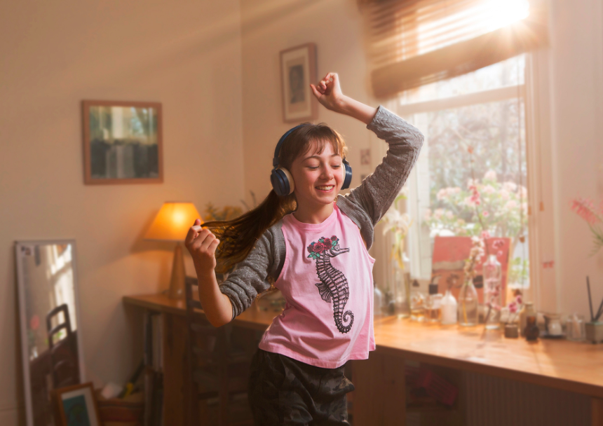 A girl with long brown hair wearing headphones dances indoors in a room with natural light streaming through the window and across the furniture.