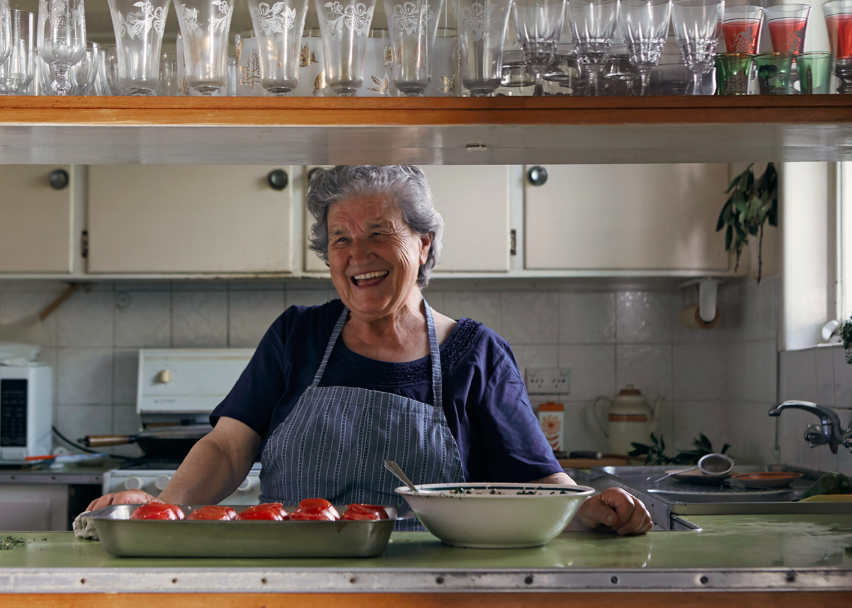 Smiling elderly woman in a kitchen setting, wearing an apron, with a tray of tomatoes and a bowl on the counter, cooking indoors.