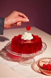A hand placing a maraschino cherry onto a fluted red gelatin dessert with whipped cream on a glass pedestal cake stand.