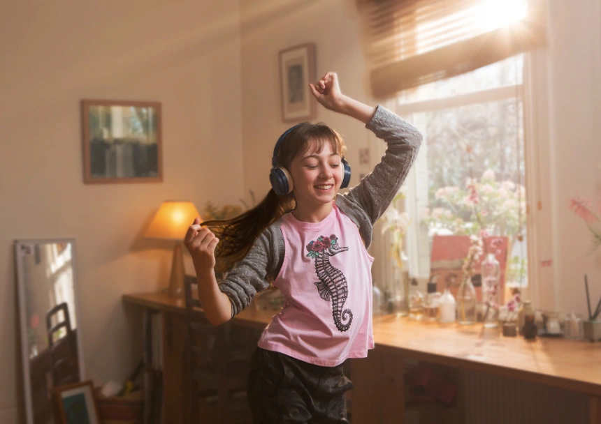 Young girl wearing pink t-shirt and headphones dancing in a sunlit bedroom by Heather Dinas