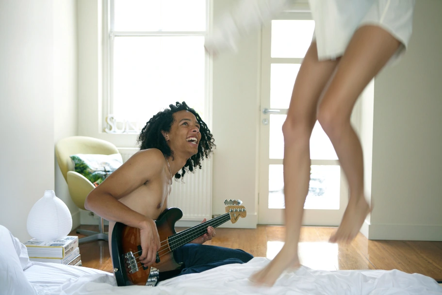 A man with dark curly hair sitting on a bed playing a bass guitar while a woman jumps nearby in a bright room.