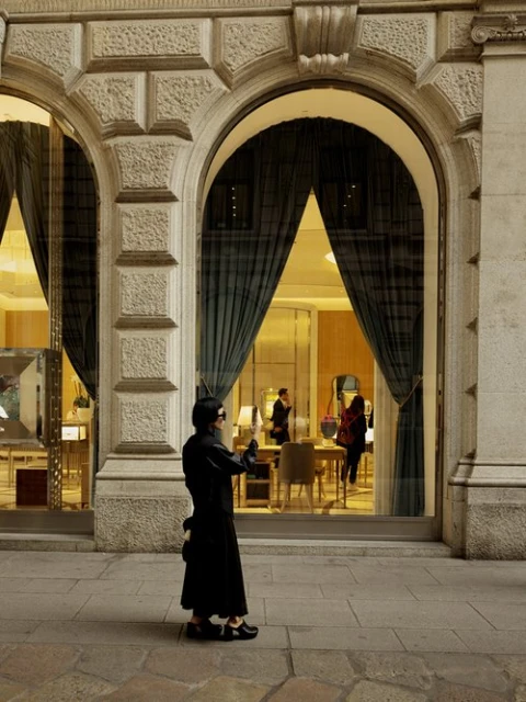 Woman in black coat stands near the arched storefront of a luxury retail shop, with interior details visible through large windows.