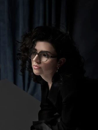 A woman with curly black hair wearing patterned glasses and a dark blazer against a dark draped backdrop.