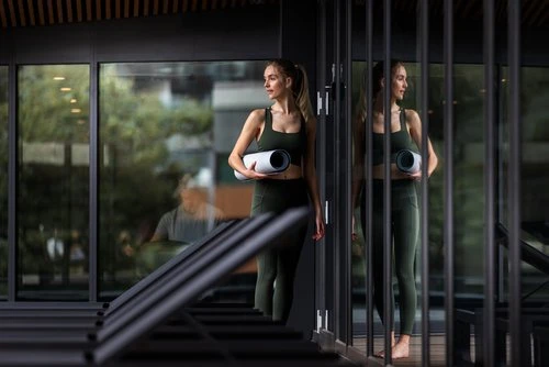 Fit woman in activewear holds a yoga mat against a windowed backdrop, bathed in natural light. Modern design with visible reflection.