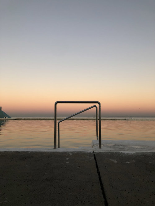 Still water in a concrete pool at dusk with metal railings and a soft color gradient in the sky from light blue to orange.