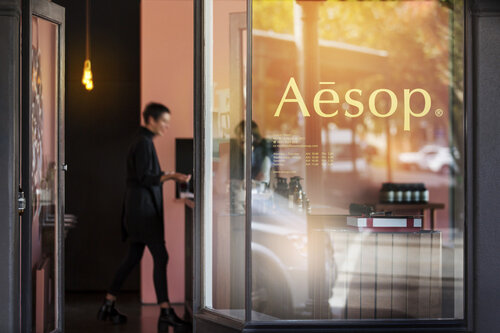 Woman entering Aesop store with window display and products in blurred background, offering skincare, haircare, and body care products.