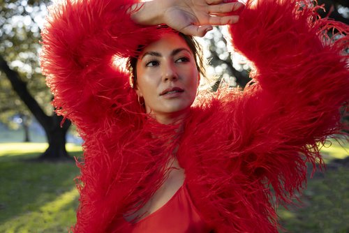 Woman in red feather coat poses outdoors in bright sunlight. Fashion shot with trees and foliage background.