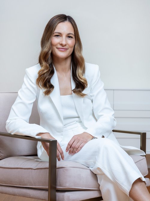 Portrait of a woman with wavy hair, wearing a white pantsuit, sitting in a mauve chair against a neutral wall in an indoor setting.