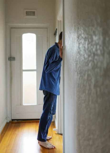 A person in a blue shirt and jeans stands facing a wall in a hallway with a white door, hardwood floors, and neutral tones, creating a sense of mystery.