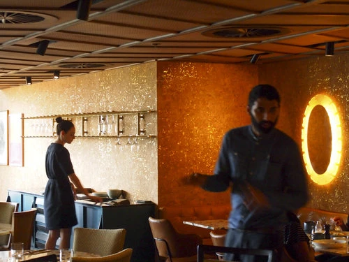 Warm-toned interior shot of two adults in a restaurant setting with gold walls, tables set for dining, and indirect overhead lighting.