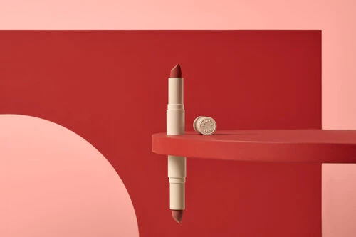 Still life of lipstick tube and cap on a red geometric platform against a pink and red background, beauty product photography.