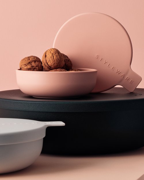 Still life shot of walnuts in a pink bowl, with a round pink paddle and a black container against a light pink background.