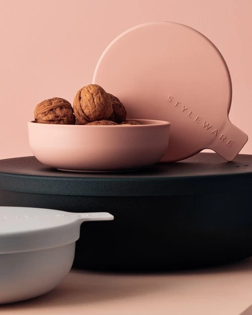 Still life shot of walnuts in a pink bowl, with a round pink paddle and a black container against a light pink background.