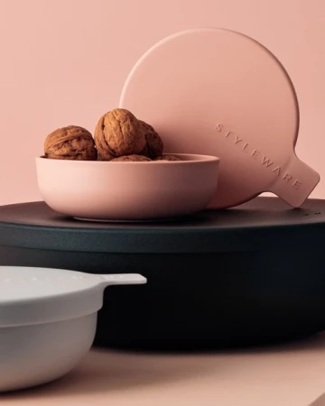 Still life shot of walnuts in a pink bowl, with a round pink paddle and a black container against a light pink background.