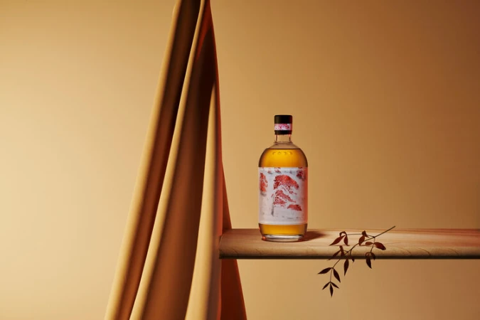 A bottle of amber liquid with a stylized label sits on a wooden surface, beside a draped peach cloth and dried leaves against a peach backdrop.