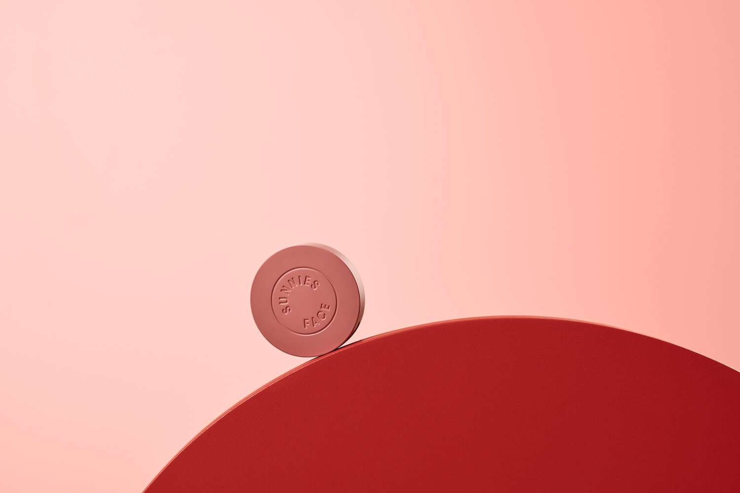 Close-up studio shot of a circular coral-colored 'Sunnies Face' product on a red platform against a gradient pink background.