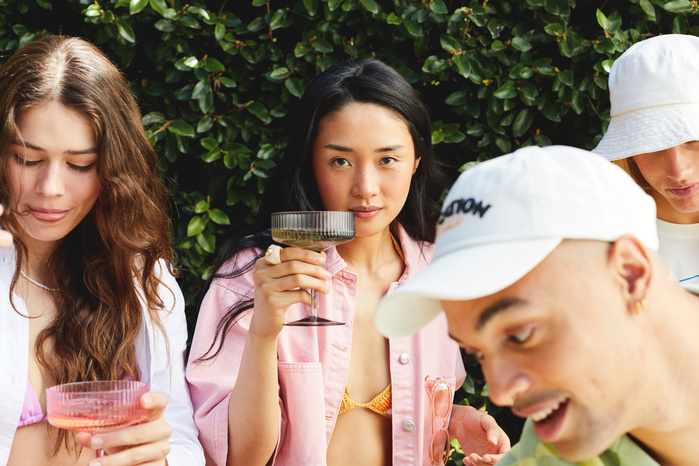 A group of young adults, including an Asian woman holding a cocktail glass, are enjoying drinks outdoors against a green foliage backdrop.