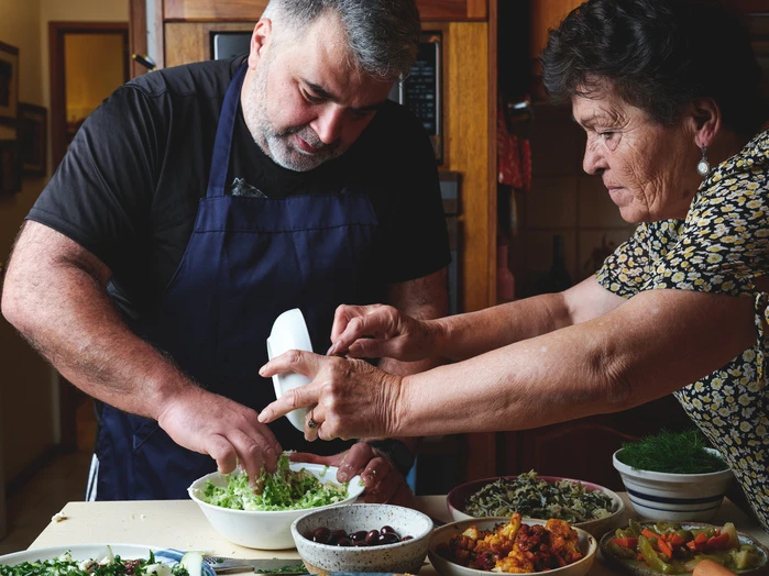 An adult male and female work together preparing food, with bowls of salad and other foods arranged on the counter in an indoor kitchen.