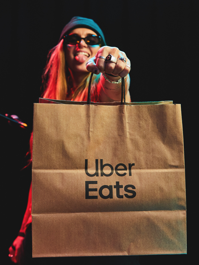 Young woman holding an Uber Eats bag in a dimly lit indoor setting with her tongue out, wearing sunglasses and a beanie.