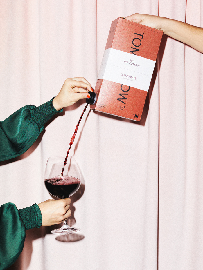 Pouring red wine from a box into a wine glass in front of a pink curtain backdrop, showcasing wine service in an indoor environment.