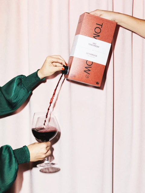 Pouring red wine from a box into a wine glass in front of a pink curtain backdrop, showcasing wine service in an indoor environment.
