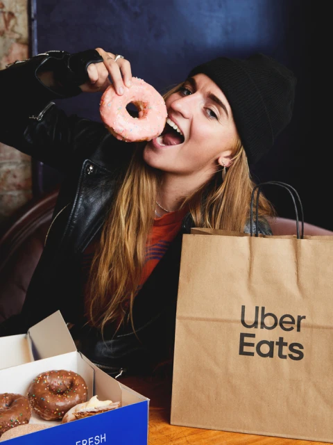 Woman in black beanie and jacket eating pink donut in indoor setting with Uber Eats bag and donut box on table.