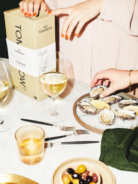 Oysters, lemon, white wine, box, forks, and olives are on a marble table with female hands reaching for oyster.