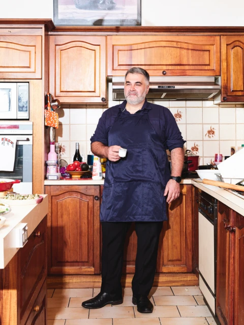 A man in a blue apron stands in a kitchen with wood cabinets, holding a cup. The image is well-lit and the focus is on the man.