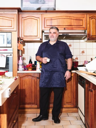 A man in a blue apron stands in a kitchen with wood cabinets, holding a cup. The image is well-lit and the focus is on the man.