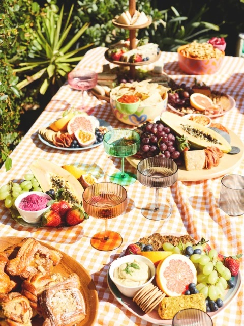 A cheerful outdoor picnic setting with colorful drinks, fresh fruit, gourmet snacks, and checkered tablecloth on a sunny day.