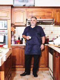 Middle-aged man wearing a dark blue apron and black trousers holding a small white cup in a traditional wooden kitchen.