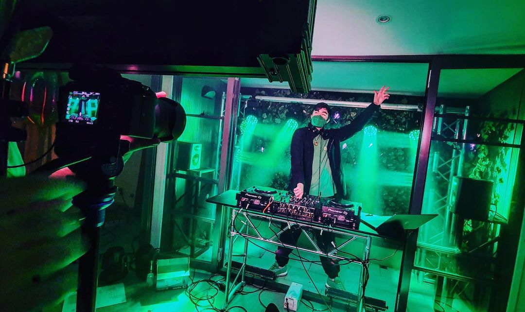 DJ performing with music equipment on stage with green lighting effects, camera recording, and masked adult