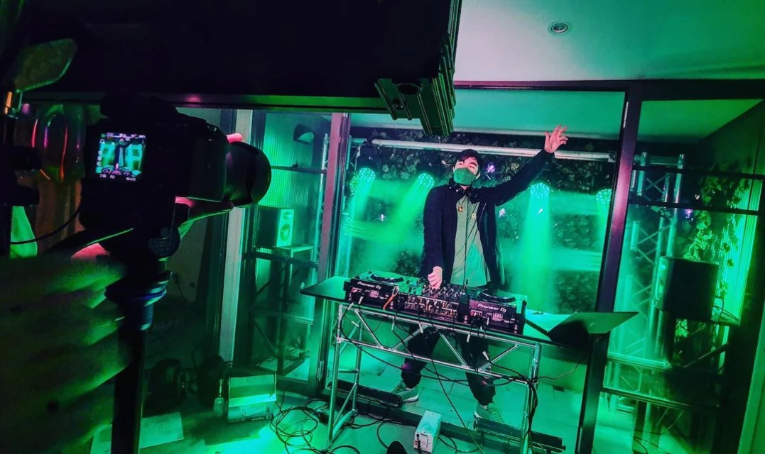 DJ performing with music equipment on stage with green lighting effects, camera recording, and masked adult