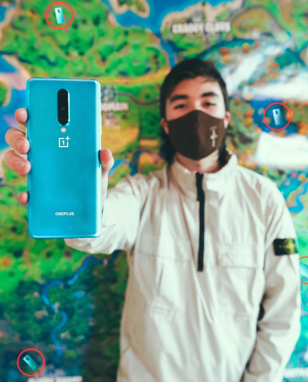 Person holds OnePlus phone with a Fortnite game map background; they're wearing a jacket and face mask in a light indoor setting.