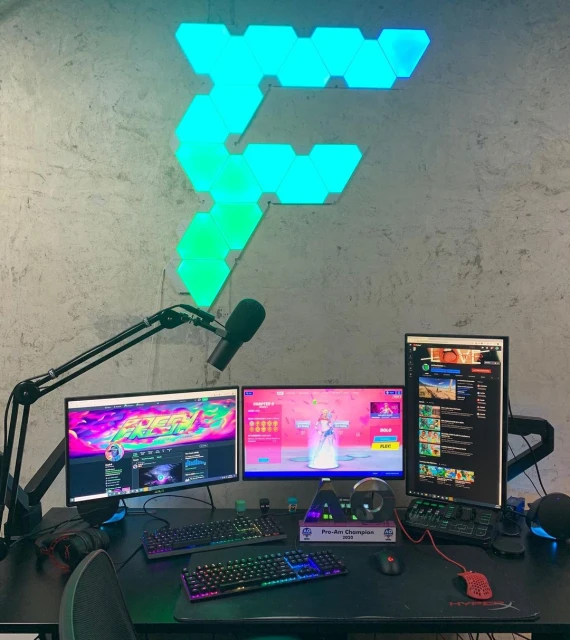 Gaming setup with three monitors, microphone, keyboard, and PC on a dark desk against a textured wall with a decorative light.