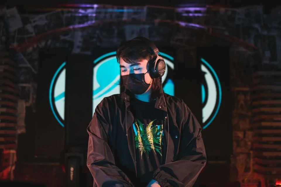 A young adult DJ wearing headphones and a black face mask stands in front of a colorful graphic background in a dimly lit club.