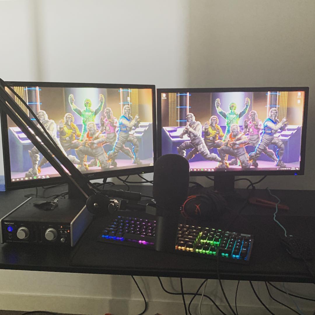 Gaming setup featuring two monitors displaying video game characters, a microphone, keyboard, and audio interface on a black desk indoors.