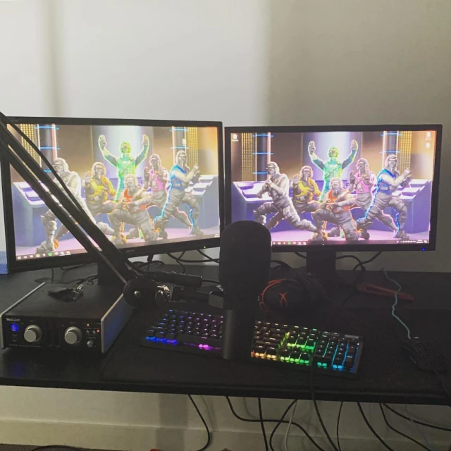 Gaming setup featuring two monitors displaying video game characters, a microphone, keyboard, and audio interface on a black desk indoors.