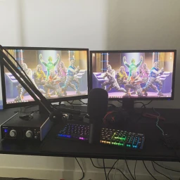 Dual computer monitors displaying video game characters on a desk with an audio interface, microphone, and RGB keyboard