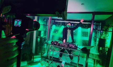 A DJ performing behind a mixing console with vibrant green stage lighting in a room while a camera films the set from the foreground.