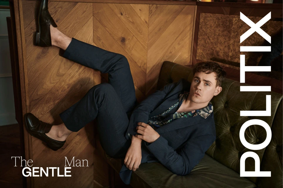 A young man poses while reclining on a green tufted sofa, wearing a blue suit and loafers, with wood paneling in the background.
