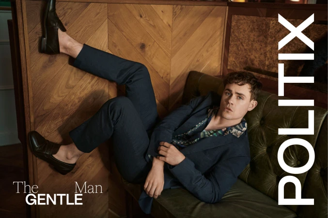 A young man poses while reclining on a green tufted sofa, wearing a blue suit and loafers, with wood paneling in the background.