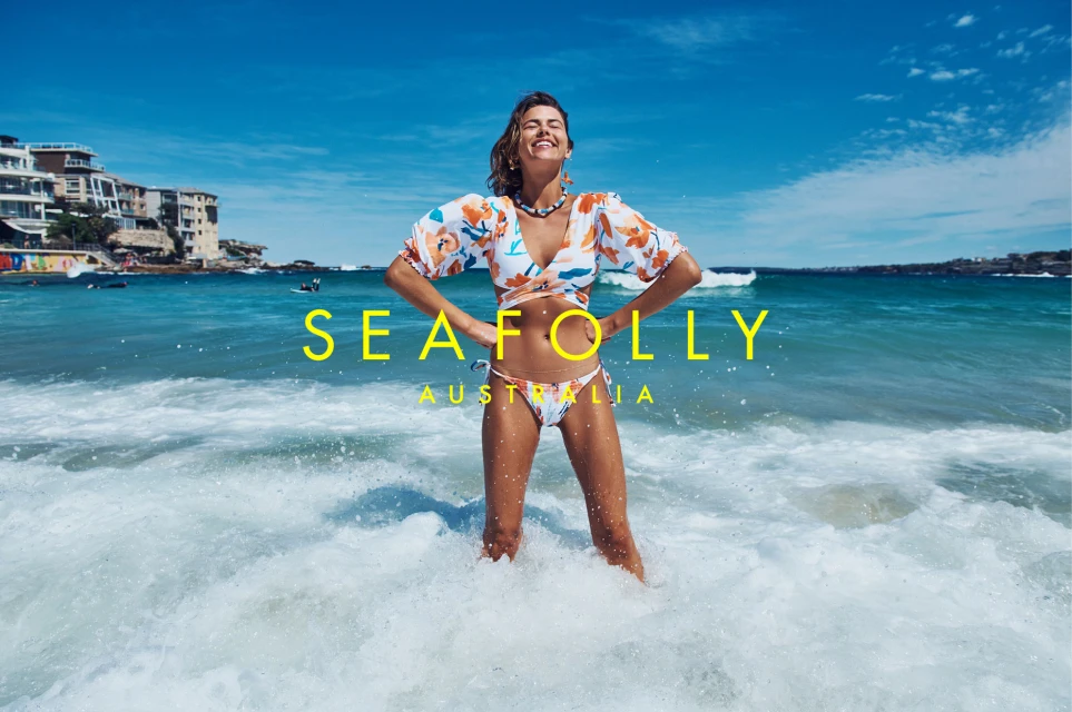 Adult woman in a floral bikini and top stands in the ocean surf on a sunny day, with buildings visible on the coast in the background.