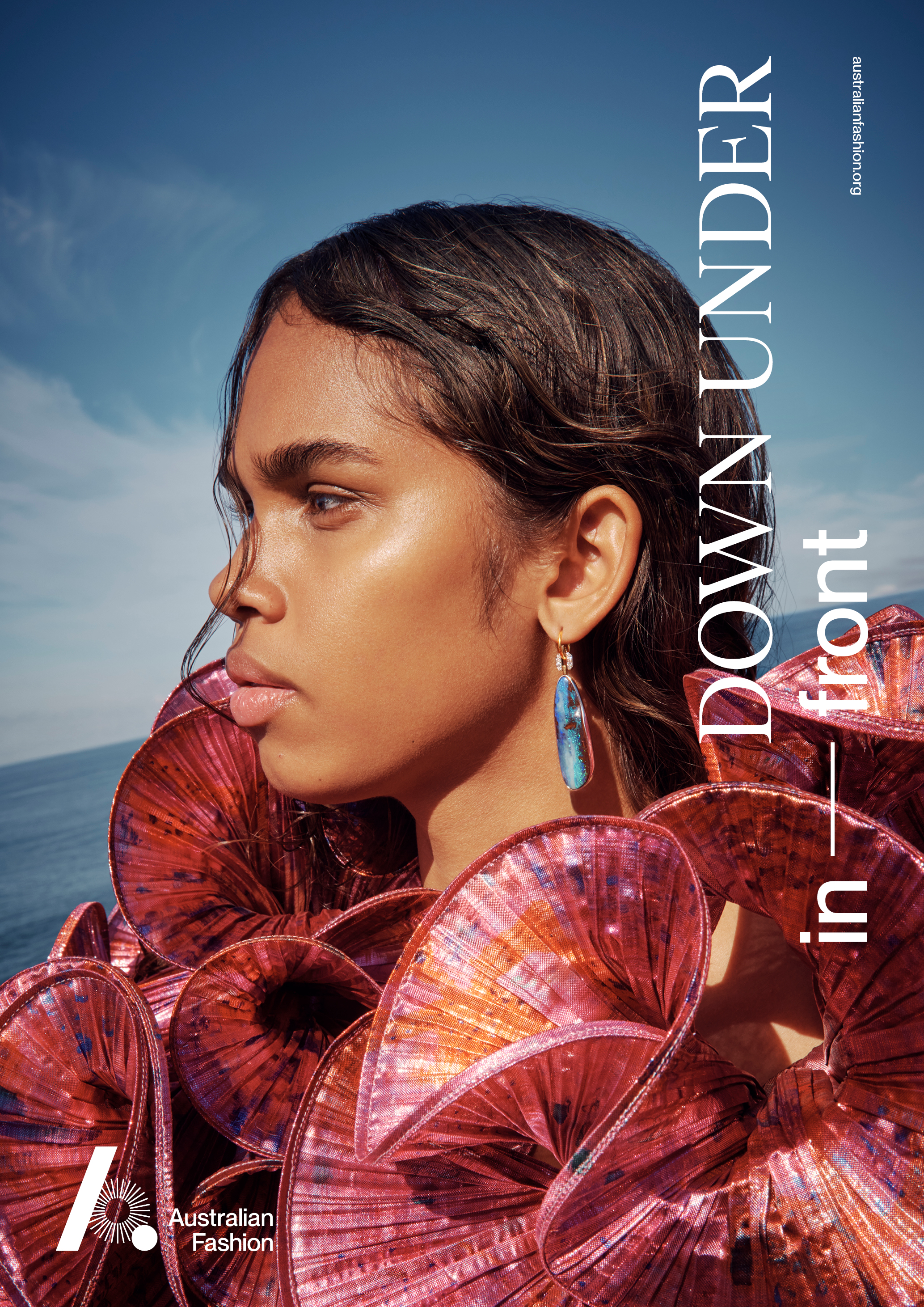 Fashion editorial shot of a woman with dark hair, wearing a sculptural red top and earring, against a blue sky and ocean backdrop.