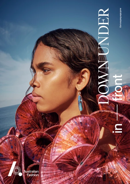 Fashion editorial shot of a woman with dark hair, wearing a sculptural red top and earring, against a blue sky and ocean backdrop.