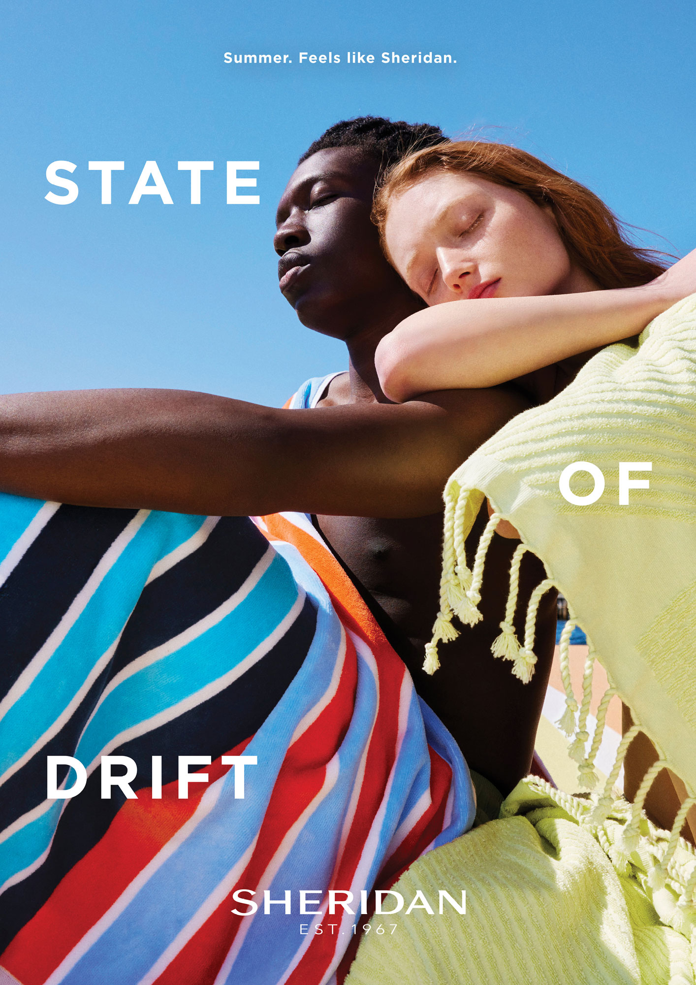 An interracial couple reclines against a brilliant blue sky. The pair is draped in colorful striped and solid colored Sheridan towels.